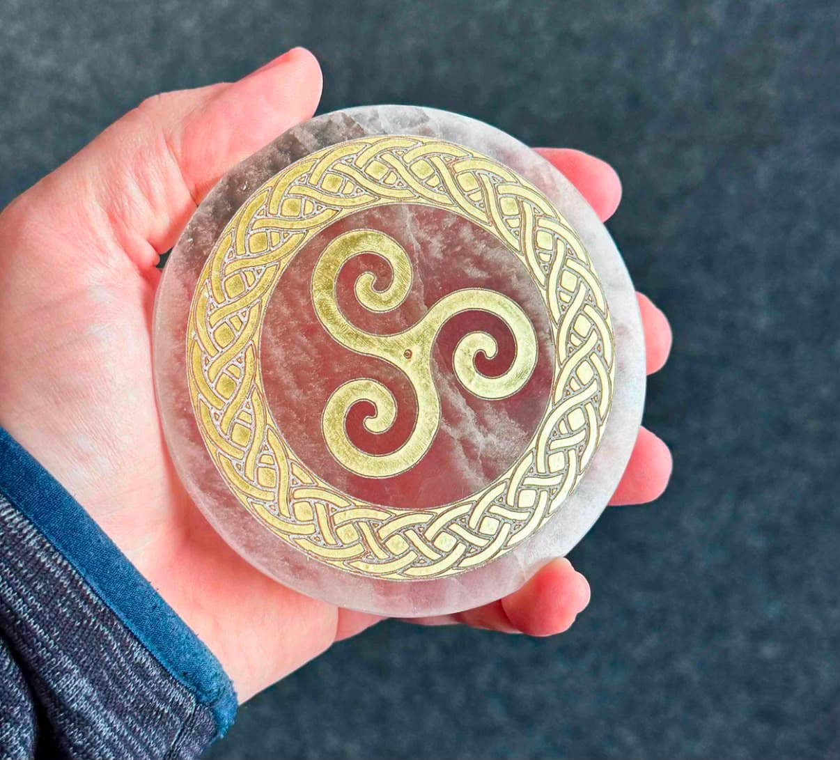 Hand holding a translucent stone disc featuring a gold triskelion and Celtic knotwork border.