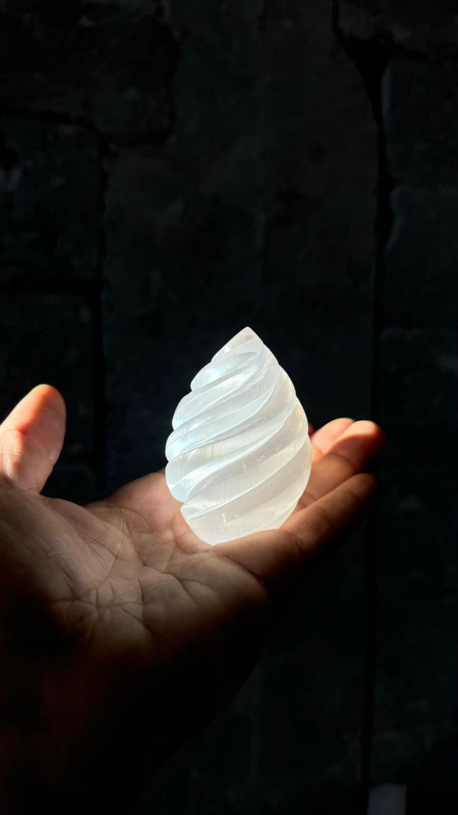 Mano sosteniendo una espiral de cristal de selenita blanca brillante sobre un fondo de piedra oscura.