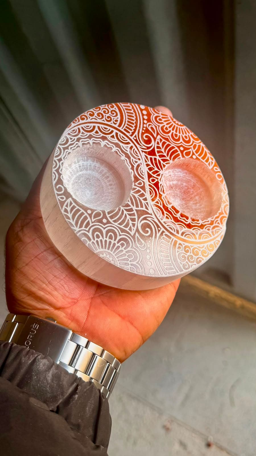 Mano sosteniendo un portavelas redondo de selenita con intrincados patrones de mandalas blancos y matices anaranjados.